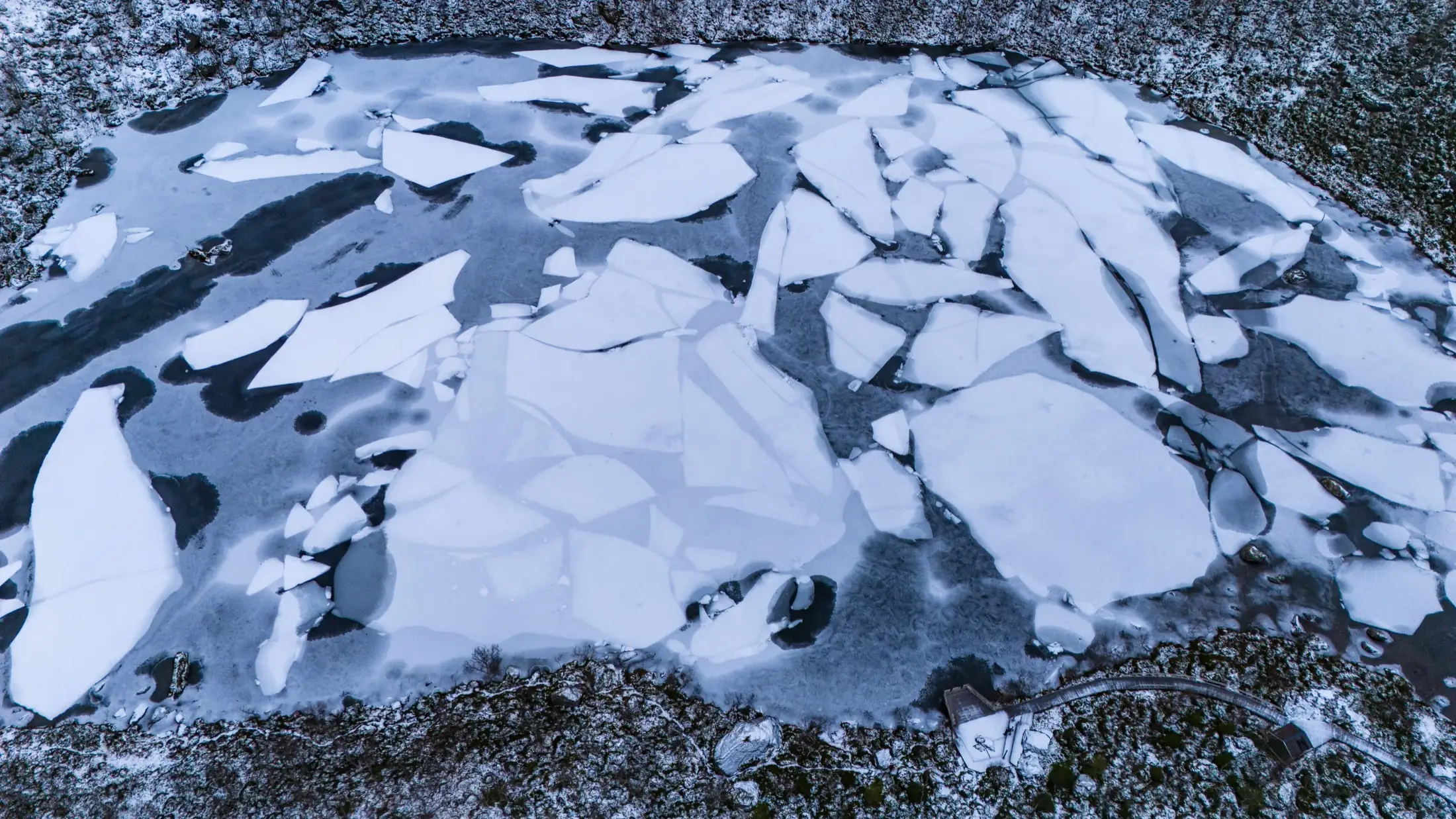 Un lac gelé vu du ciel, ses grandes plaques de glace et la forêt enneigée sur les rives.